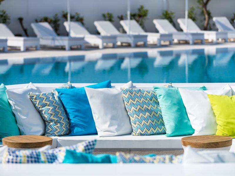 Kolorowe poduszki na leżance obok spokojnego basenu hotelowego z leżakami w tle.