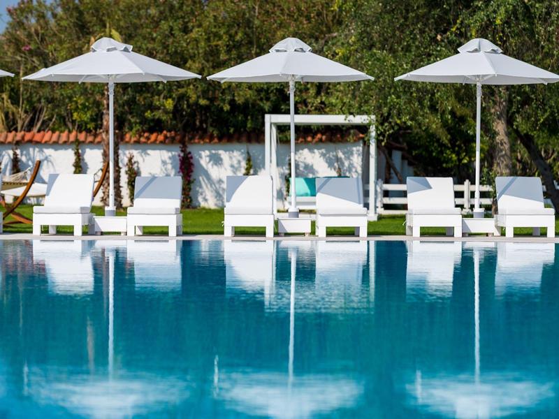 Widok na rząd białych leżaków i parasoli obok czystego niebieskiego basenu z zielonym krajobrazem w tle.