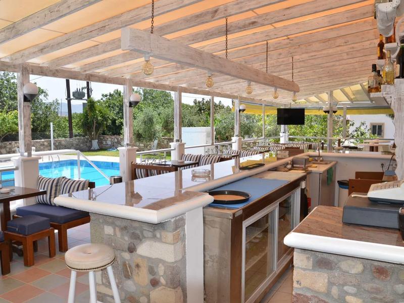 Otevřený bar s výhledem na bazén a zahradu, světlý dřevěný strop a posezení.