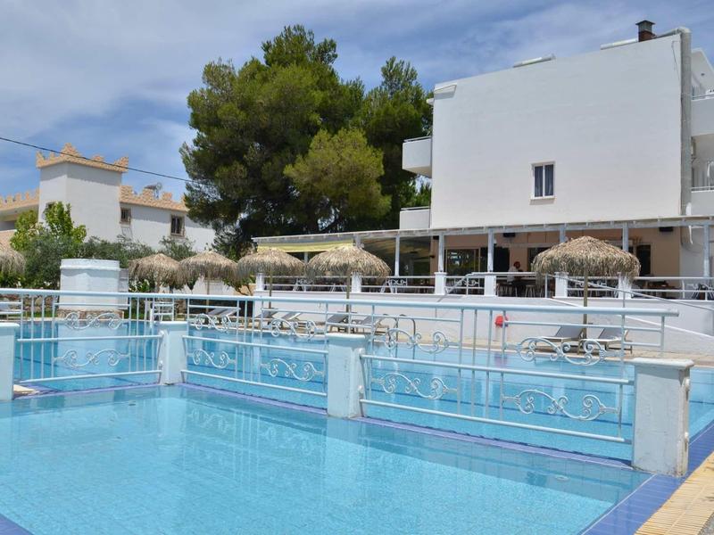 Moderní venkovní bazén s slunečníky před bílou hotelovou budovou pod modrou oblohou.