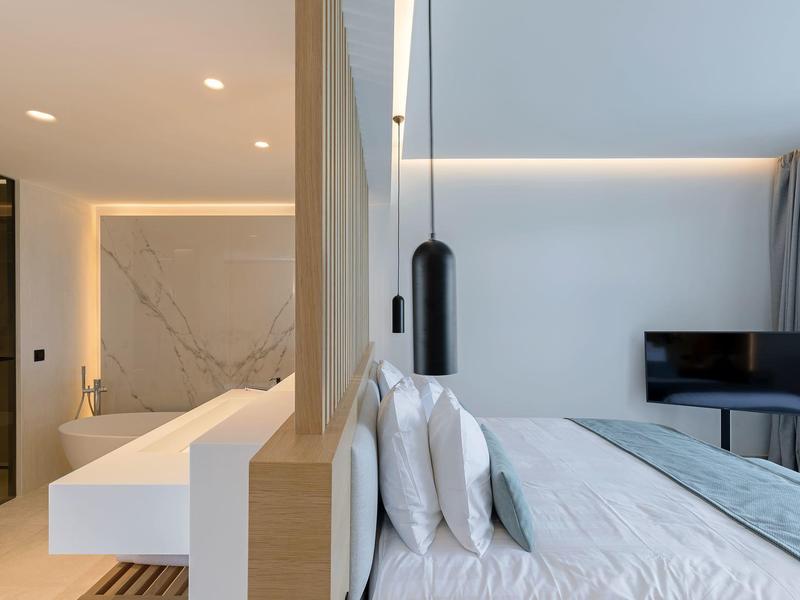 Modern hotel room with white bed, black lamp, and TV against light-colored walls.