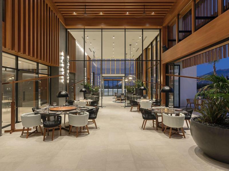 Modern hotel dining area with wooden ceiling, large plants, and comfortable chairs in the evening.