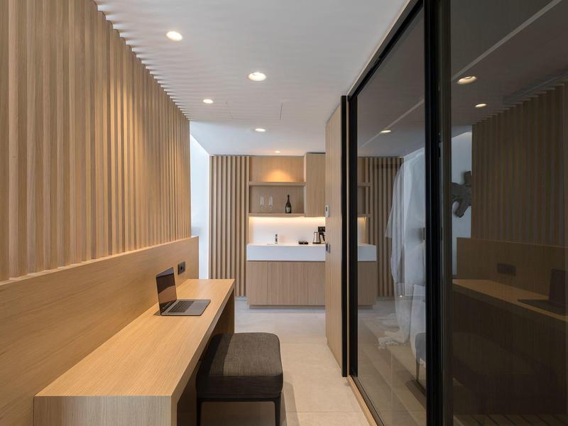 Modern hotel room workspace with wood paneling, desk, and adjoining bathroom separated by glass wall.