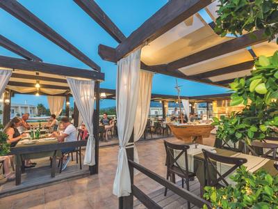 Außenrestaurant mit Holzbalken, weißen Vorhängen, Tischen und Stühlen bei Abenddämmerung.
