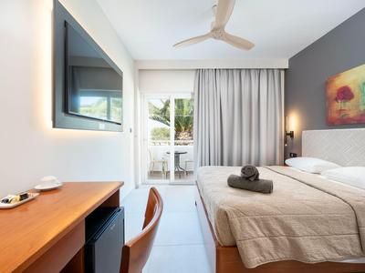 Helles Hotelzimmer mit Doppelbett, Schreibtisch, Fernseher und Zugang zu Balkon mit Gartenblick.