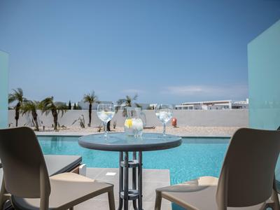 Terrasse mit Stühlen und kleinem Tisch am Pool in einer Hotelanlage bei blauem Himmel.