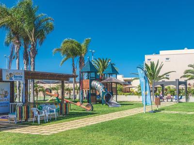 Grüner Garten mit Palmen, Spielplatz und weißem Gebäude unter strahlend blauem Himmel.