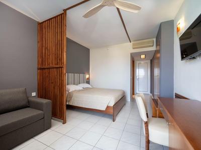 Modernes Schlafzimmer mit weißem Bett, grauem Sofa, Holzwand, Fernseher und Schreibtisch.