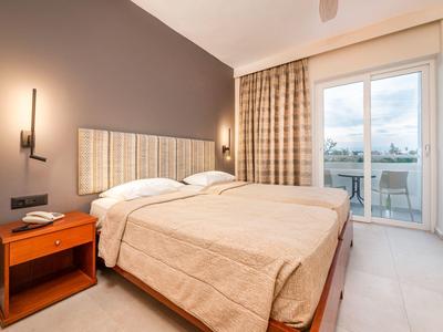 Schlafzimmer mit Doppelbett, beige Bettdecke, Holz-Nachttisch und großem Fenster mit Vorhängen.