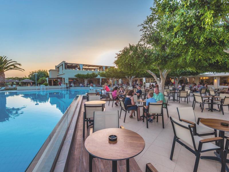 Área externa de un restaurante junto a una gran piscina al atardecer con clientes.