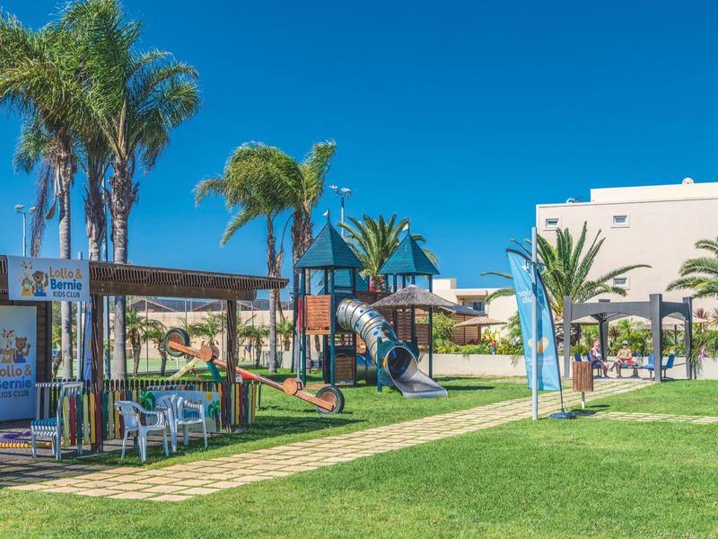 Área de juegos con toboganes, mesa y sillas frente a edificios blancos bajo cielo azul.