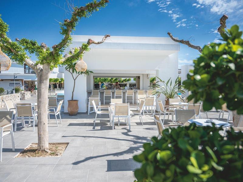 Terraza luminosa con edificio blanco, sillas y mesas bajo cielo azul y árboles.