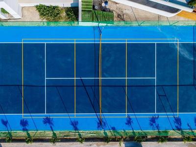 Draufsicht auf hellblaues Tennisfeld mit Netz und weißen Linien, umgeben von grünem Rasen und Gebäuden.