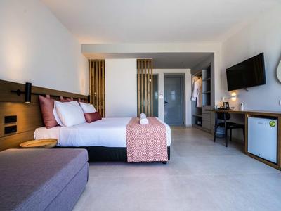 Modernes Hotelzimmer mit großem Bett, grauem Sofa, Wandfernseher und Holzmöbeln.