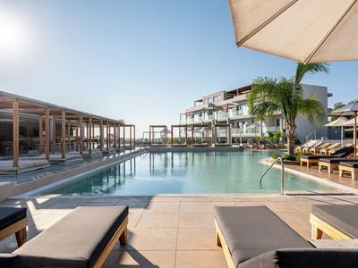 Moderner Hotelpoolbereich mit Sonnenliegen, Sonnenschirmen und Palmen bei klarem Himmel.