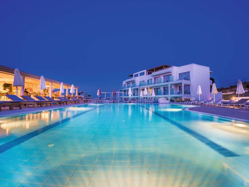 Resortzwembad bij schemering met helder water, ligstoelen en moderne witte gebouwen onder een blauwe lucht.