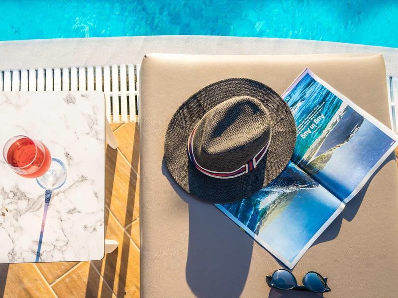 Zijaanzicht bij het zwembad met zonnehoed, zonnebril, tijdschrift en drankje op een ligstoel.