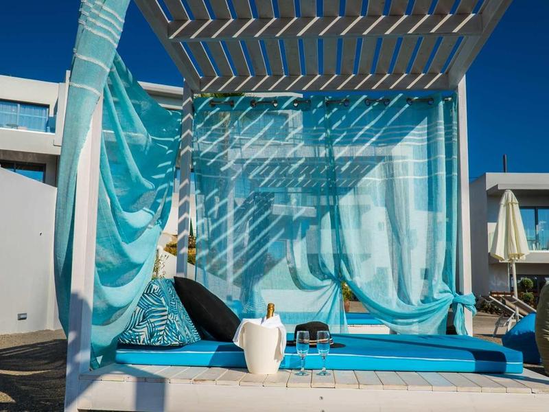 Buitenloungebed met blauwe gordijnen en luifel bij een zonnig resort zwembadgebied.