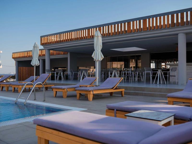 Moderne hotelzwembad met ligbedden, parasols en bar onder een heldere blauwe lucht.