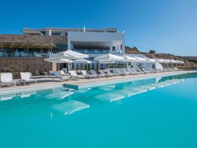 Moderní hotelový komplex s velkým bazénem a bílými slunečníky pod jasně modrou oblohou.