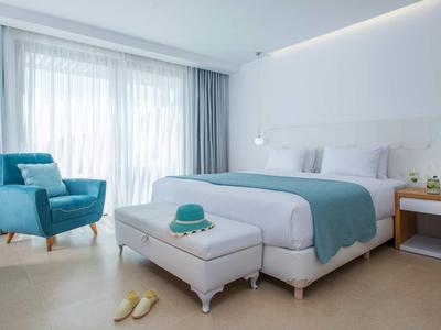 Nowoczesny pokój hotelowy z dużym łóżkiem, niebieskim fotelem i światłem dziennym przez duże okna.