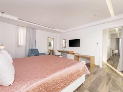 Jasny pokój hotelowy z dużym łóżkiem, różowym narzutem, miejscem do siedzenia i telewizorem na ścianie.