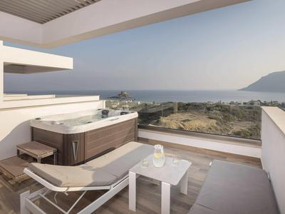 Balkon z jacuzzi, leżakiem i stolikiem z widokiem na morze i góry w pogodny dzień.