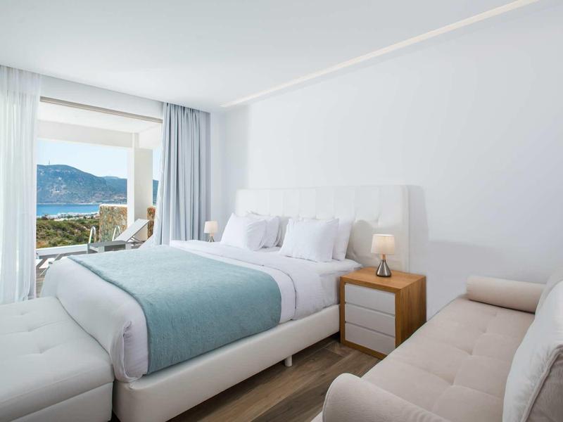 Jasny pokój hotelowy z łóżkiem podwójnym, sofą i balkonem z widokiem na morze.