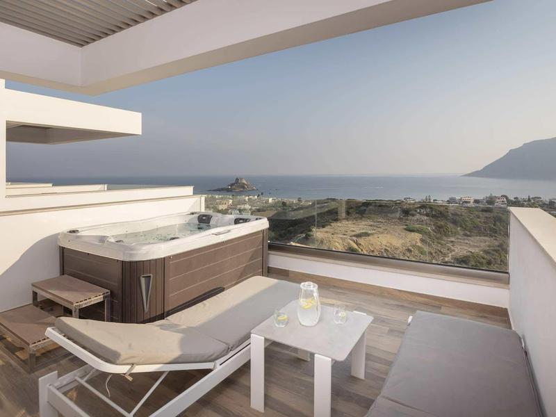 Balkon z jacuzzi, leżakiem i stolikiem z widokiem na morze i góry w pogodny dzień.