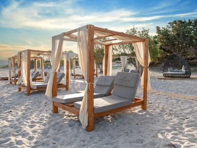 Pláž s dřevěnými postelemi s baldachýnem a polštáři, klidný západ slunce a stromy v pozadí.