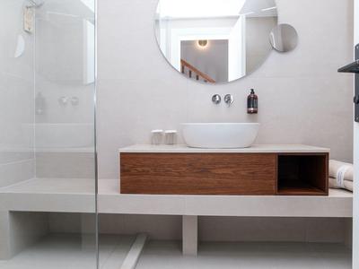 Moderní koupelna s dřevěnou skříňkou, bílým umyvadlem, kulatým zrcadlem a skleněnou sprchou.