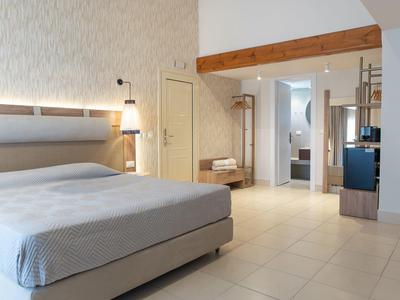 Světle osvětlený hotelový pokoj s manželskou postelí, dřevěnými trámy a světlou dlažbou.