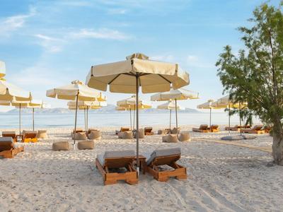 Sandstrand mit Sonnenliegen und Sonnenschirmen neben einem Baum unter blauem Himmel.
