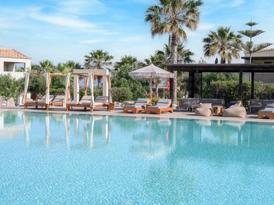 Großer Hotelpool mit Sonnenliegen, Palmen und klarem Himmel im Hintergrund.