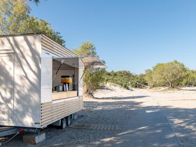 Kleines Holzferienhaus auf Sand mit Bäumen und blauem Himmel im Hintergrund.