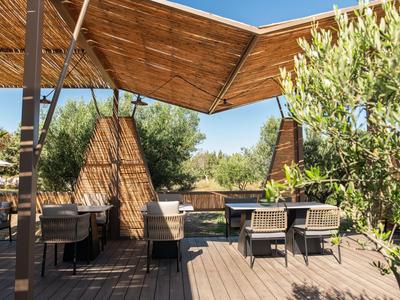 Überdachte Holzterrasse mit Tischen und Stühlen, umgeben von Pflanzen und blauen Himmel.