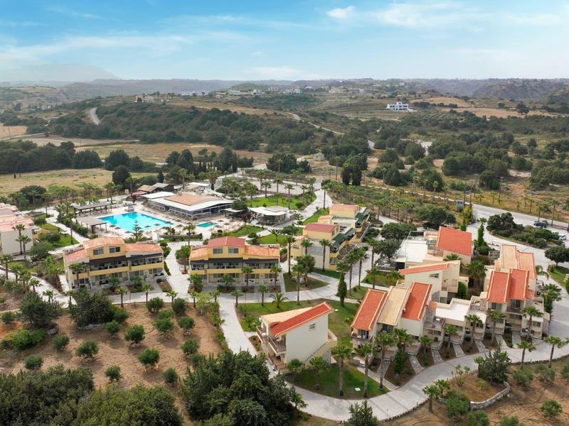 Resort s více budovami a bazény obklopený stromy a kopci pod modrou oblohou.