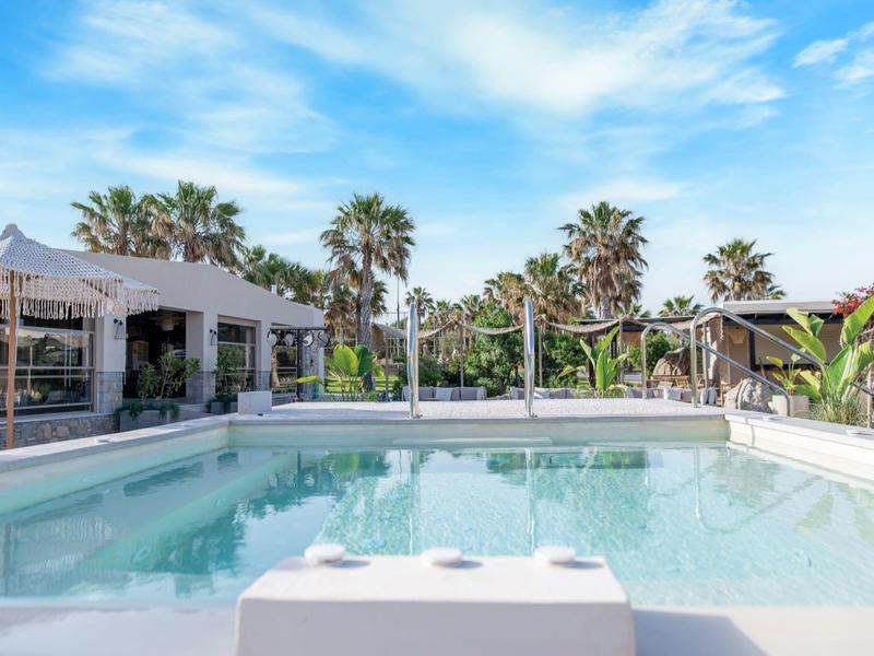 Venkovní bazén s čistou vodou obklopený palmami a moderními budovami pod modrou oblohou.