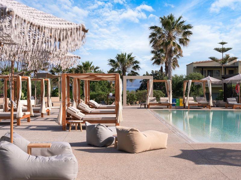 Rezortní bazénová oblast s lehátky, slunečníky, palmami a jasnou modrou oblohou.