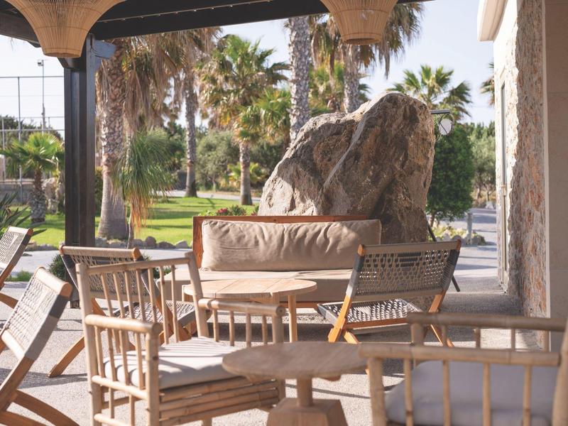 Pohodlná venkovní posezení se dřevěnými židlemi a stolem pod zastíněnou verandou poblíž palm.