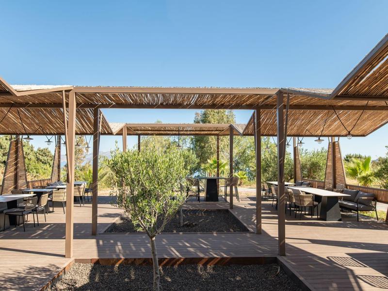 Venkovní jídelní prostor s dřevěnými pergolami a kovovými židlemi pod jasnou modrou oblohou.