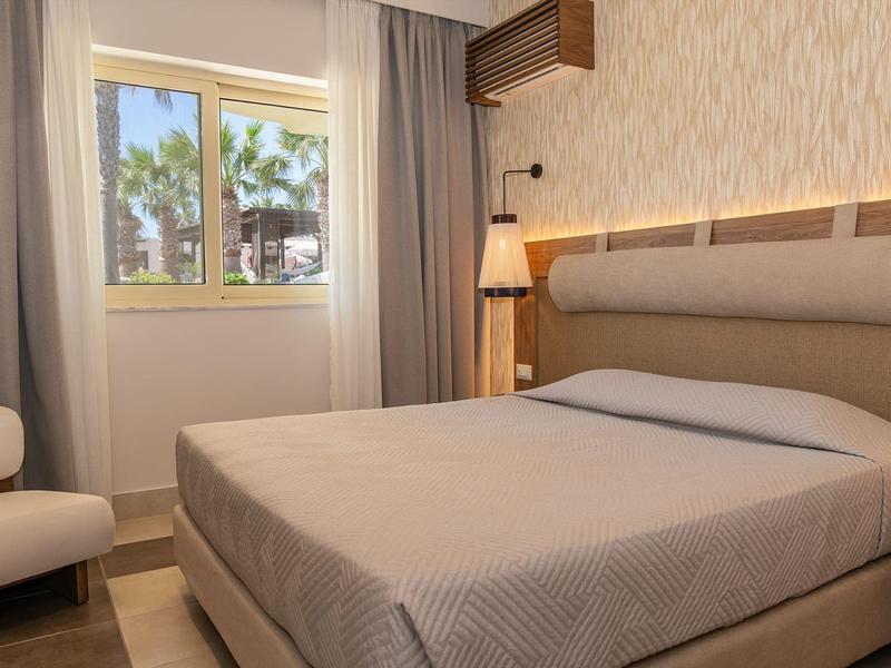 Pohodlný hotelový pokoj s velkou postelí, neutrálním zařízením a oknem s výhledem na palmy.