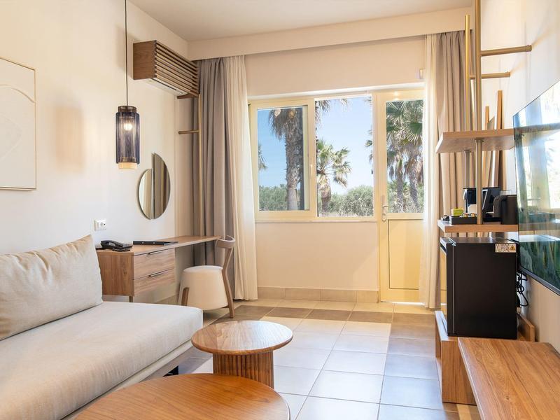 Jasný hotelový pokoj s pohovkou, dřevěnými stolky a velkým oknem s závěsy