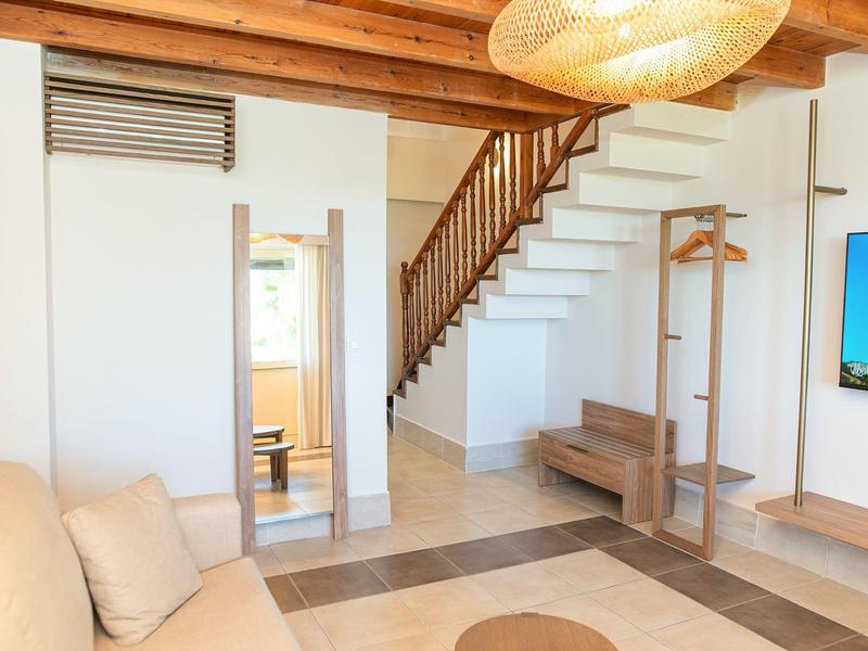 Světý hotelový pokoj s dřevěnými trámy, schodištěm, béžovou pohovkou a zavěšenou TV