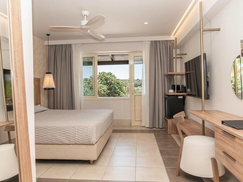 Světý hotelový pokoj s postelí, televizí, psacím stolem, židlí a velkým oknem s výhledem na zeleň.