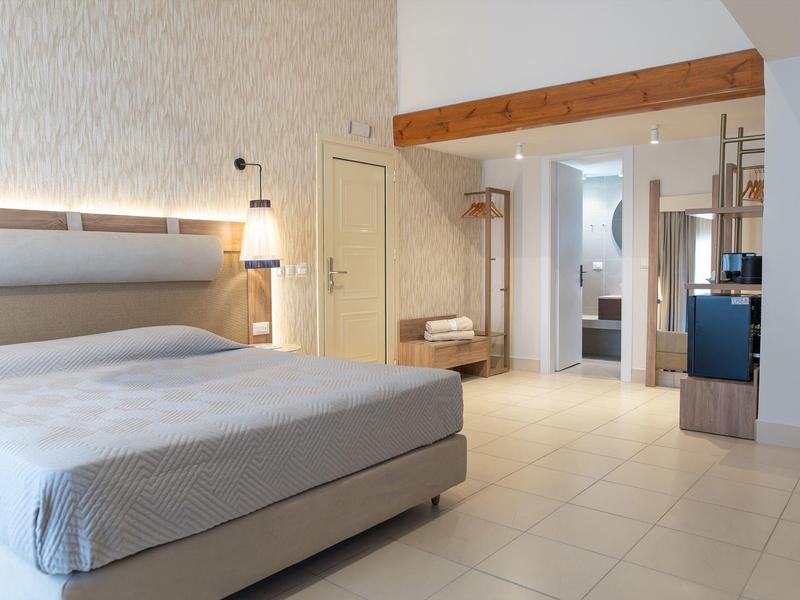 Světle osvětlený hotelový pokoj s manželskou postelí, dřevěnými trámy a světlou dlažbou.