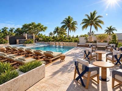 Sonniger Hotelpoolbereich mit Liegestühlen und Sitzgelegenheiten, Palmen und blauem Himmel.