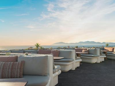 Lounge areas with comfortable sofas and cushions on a rooftop terrace overlooking the sea and mountains in the background.