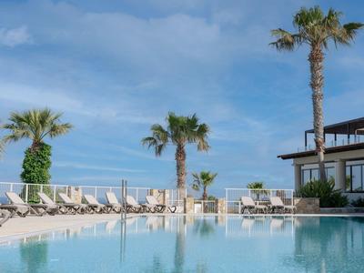 Modern hotel pool with loungers and palm trees under a blue sky by the sea.