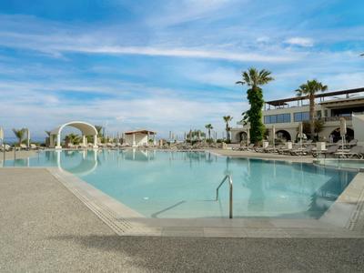 Large outdoor pool with clear water, surrounded by lounge chairs and palm trees under blue sky.